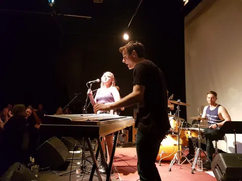 A singer at a keyboard under one spotlight, drummer behind